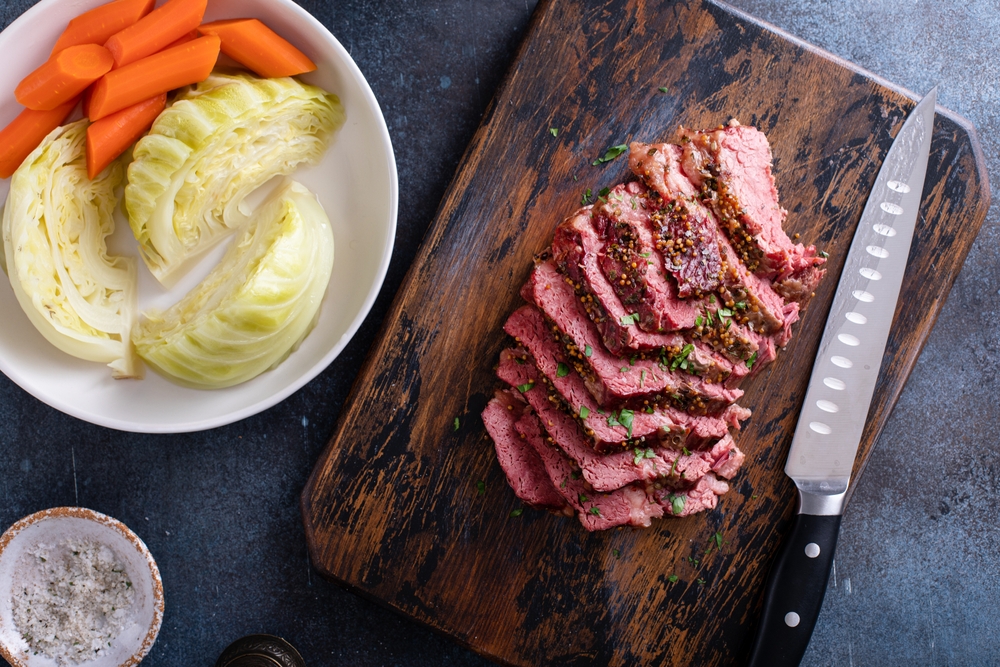 Corned beef sliced up on a cutting board ready to be added to any dish. On the table is a bowl of cabbages and carrots to add to the meal.