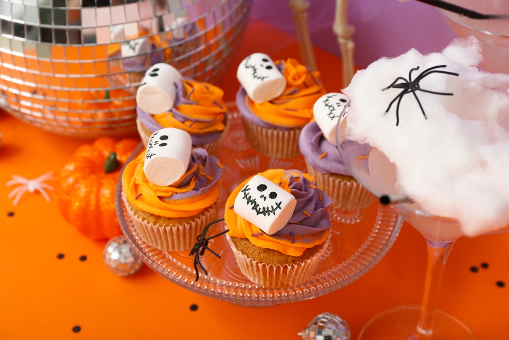 Marshmellows with faces on it, on top of orange and purple covered cupcakes.