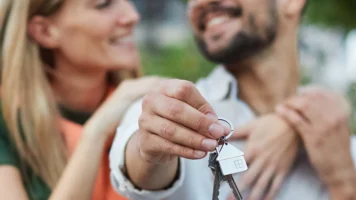 Husband and wife smile at each other while holding out keys to their new home | Things to Consider Before Refinancing Your Home Loan