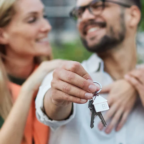 Husband and wife smile at each other while holding out keys to their new home | Things to Consider Before Refinancing Your Home Loan