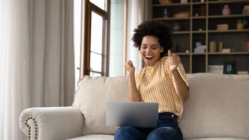 A woman grins and dances looking at her laptop, celebrating an instant approval loan