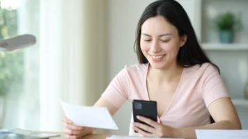Smiling woman sits at a table holding her phone and a slip of paper as she works out how to fix her credit rating