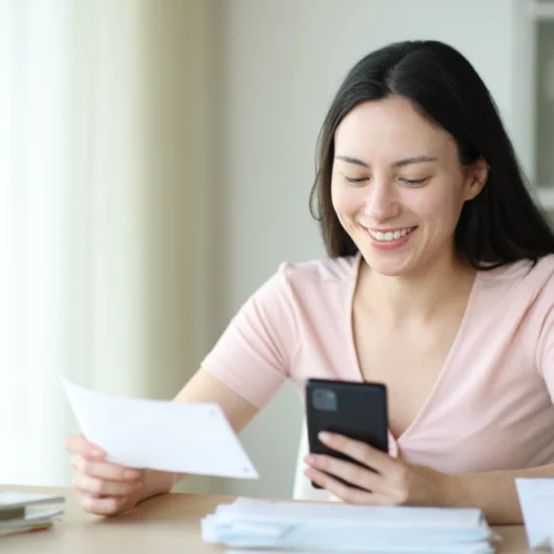 Smiling woman sits at a table holding her phone and a slip of paper as she works out how to fix her credit rating