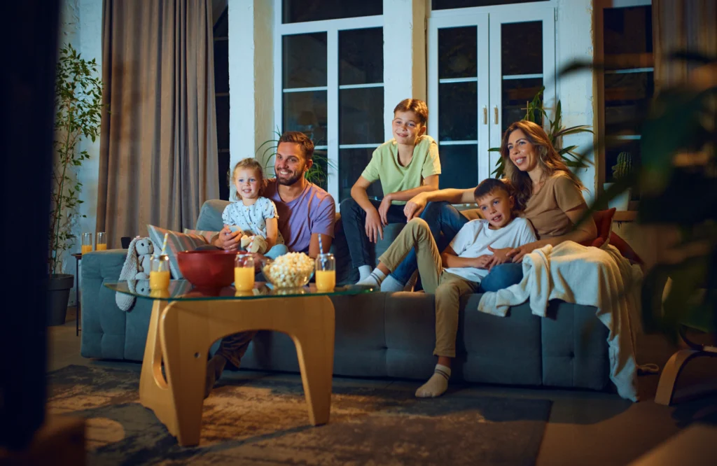 Two parents and their three kids sit on the couch with popcorn and orange juice preparing for a movie night