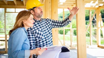 An engineer in a hard hat talks his client through the home that they're building while standing inside the wooden frame