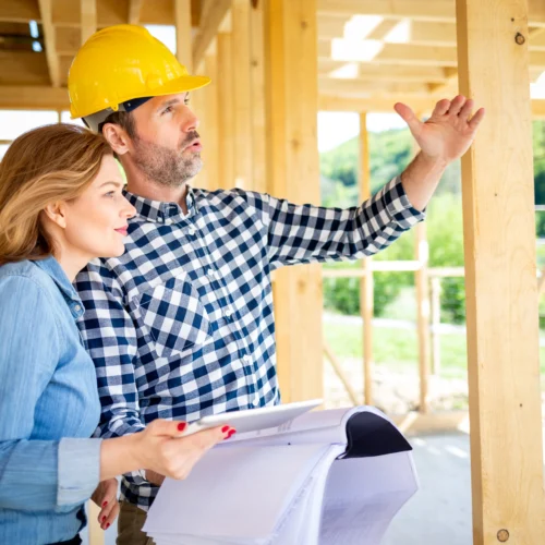 An engineer in a hard hat talks his client through the home that they're building while standing inside the wooden frame