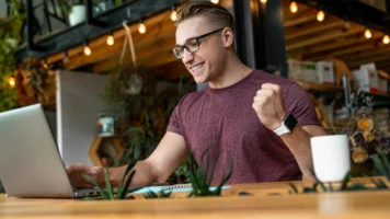 Man on his laptop in a cafe pumps his fist after getting an urgent loan with no credit check