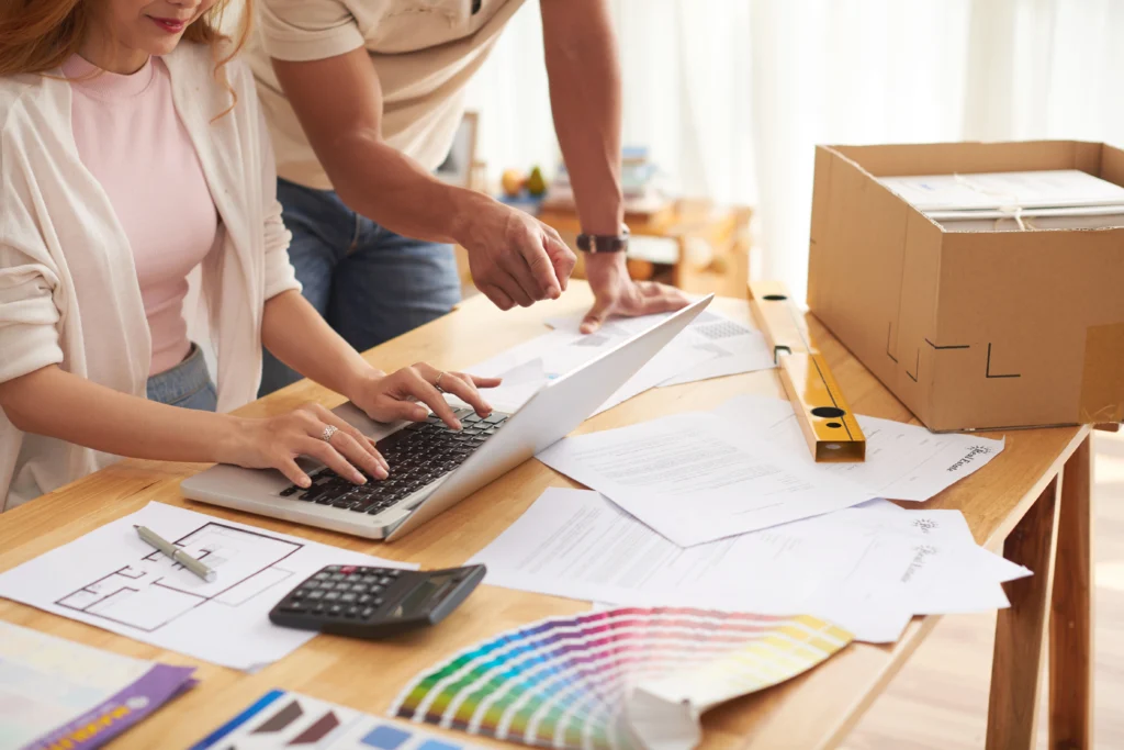 A couple at a desk full of colour swatches, building plans, and budgets looking for unexpected renovation costs