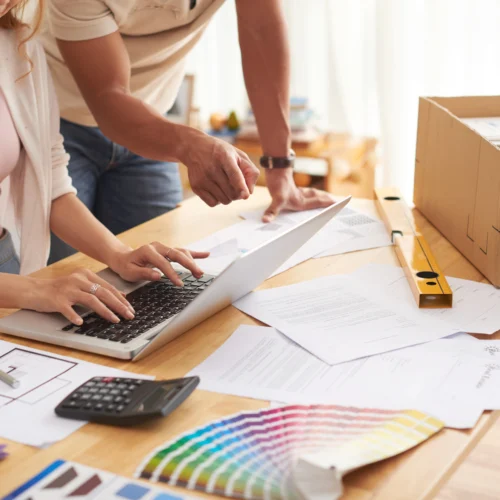 A couple at a desk full of colour swatches, building plans, and budgets looking for unexpected renovation costs