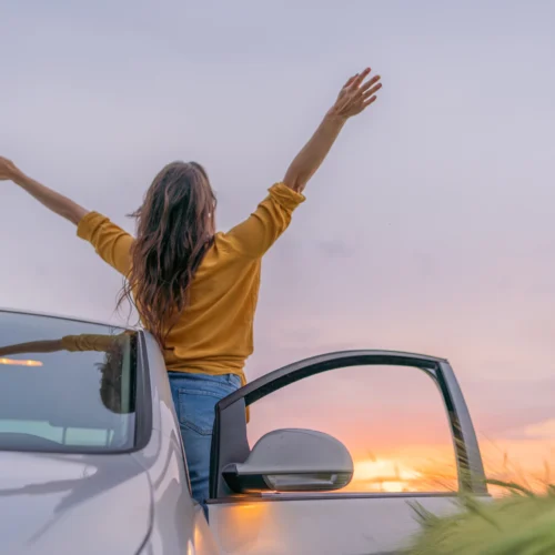 A woman leans out the drivers side of her car facing the sunset with her hands in the air