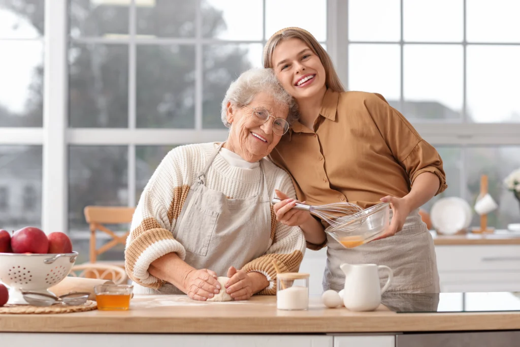 grandaughter and grandmother posing happily in kitchen kneading dough and whisking eggs as they bake together for a last minute Mother's Day gift
