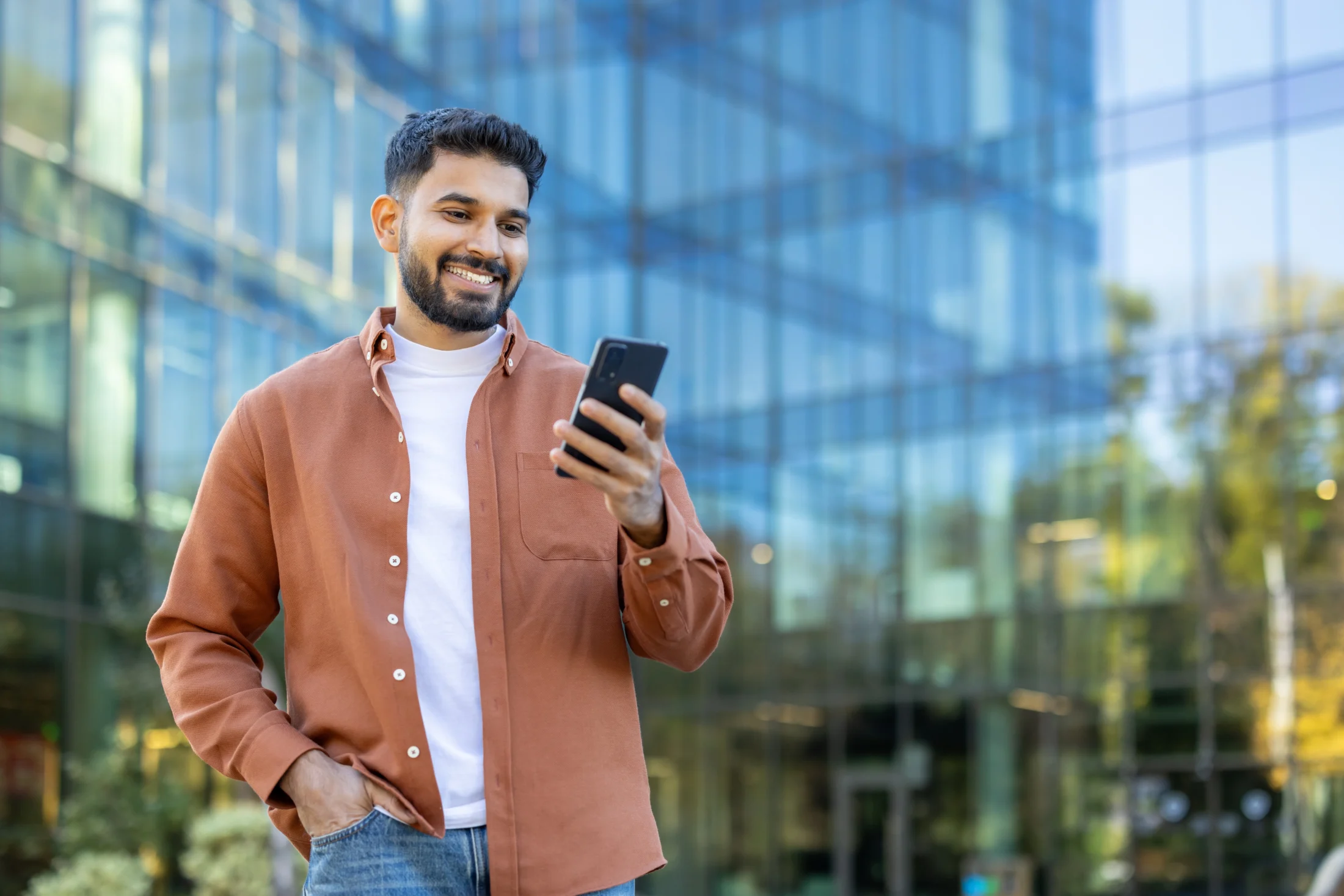 A man stands outside a modern glass building, smiling as he looks at his smartphone. He wears a brown shirt and jeans, conveying a casual, happy mood.