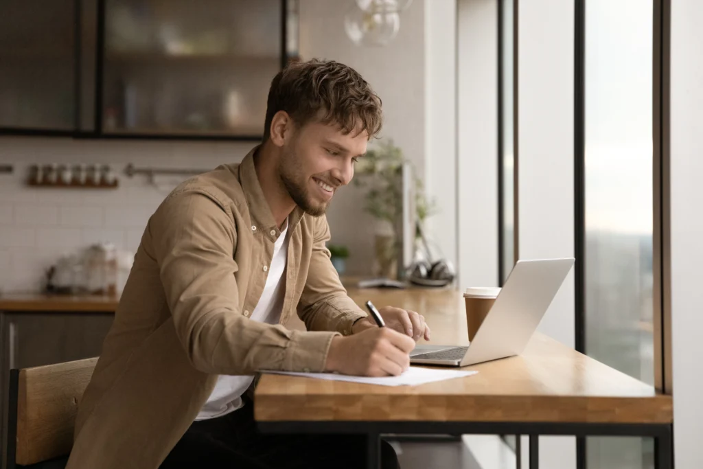 A man sits smiling at a wooden table, writing in a notebook next to an open laptop and coffee cup. Sunlight filters in through large windows, creating a warm, productive atmosphere.