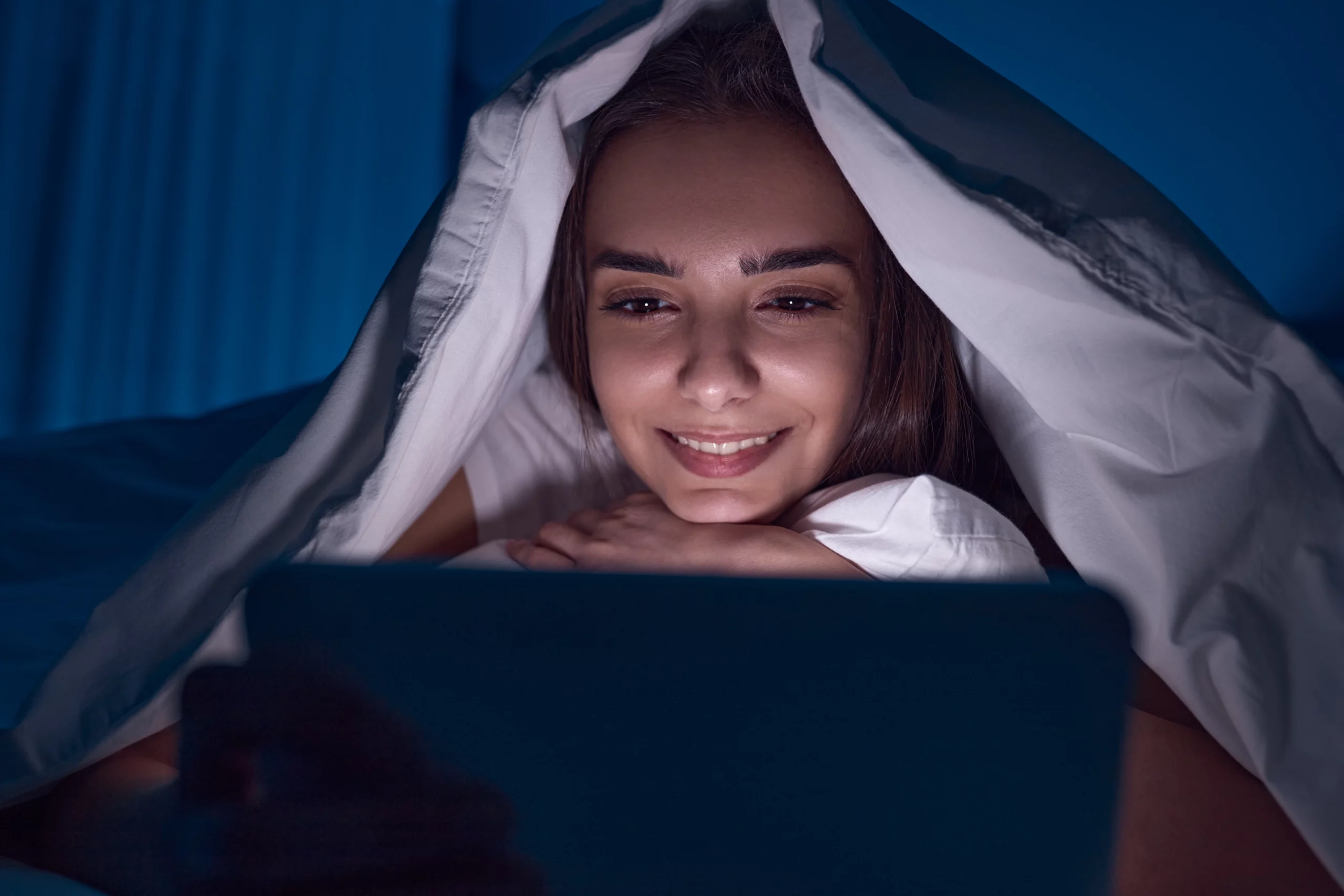 woman with blanket over her head watching a free streaming service on her laptop with the screen lighting up her face