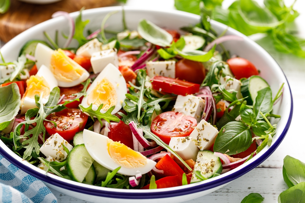 A vibrant but simple salad in a white bowl with arugula, cherry tomatoes, cucumber slices, red onion, feta, and quartered boiled eggs, garnished with herbs, conveying freshness.s