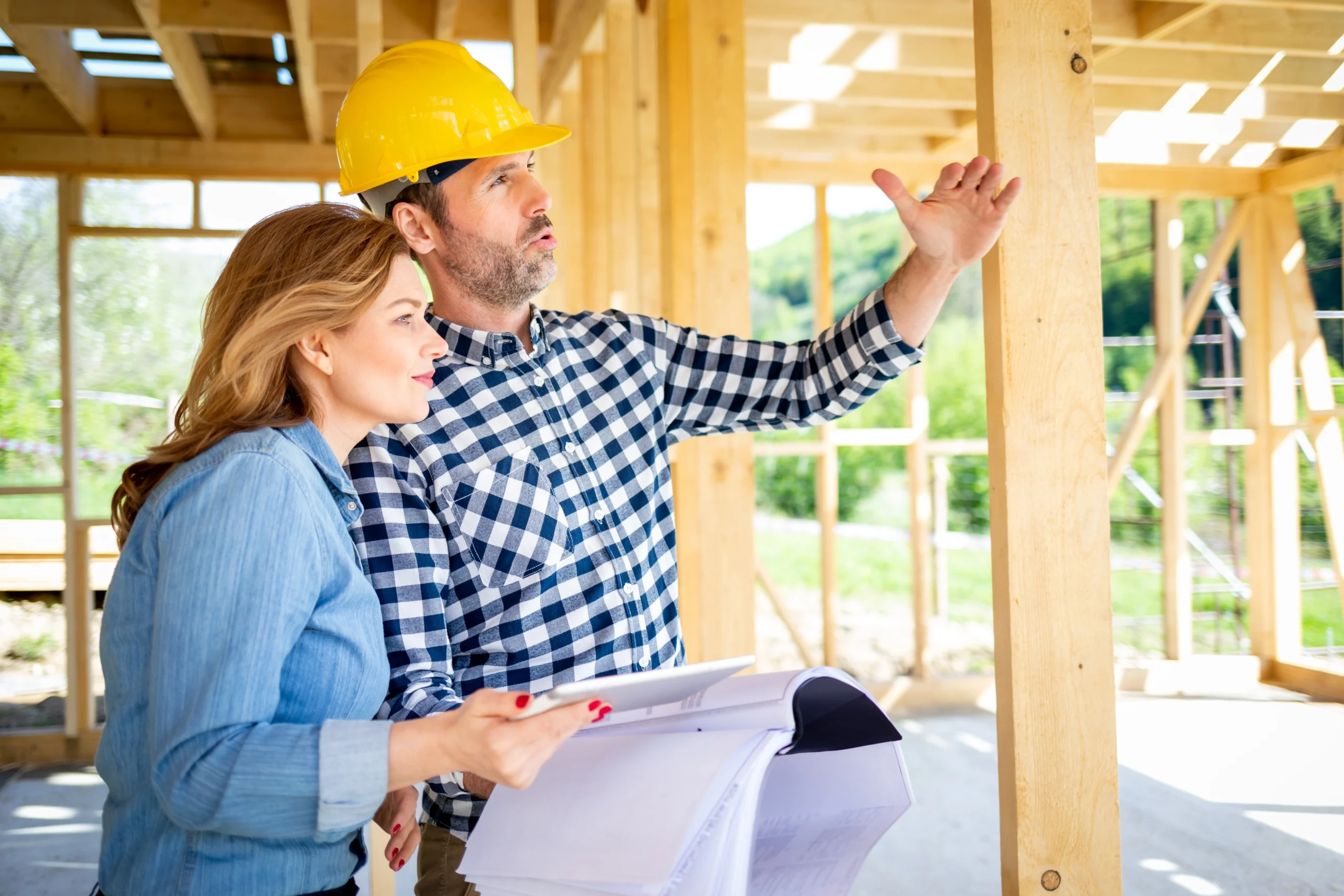 An engineer in a hard hat talks his client through the home that they're building while standing inside the wooden frame