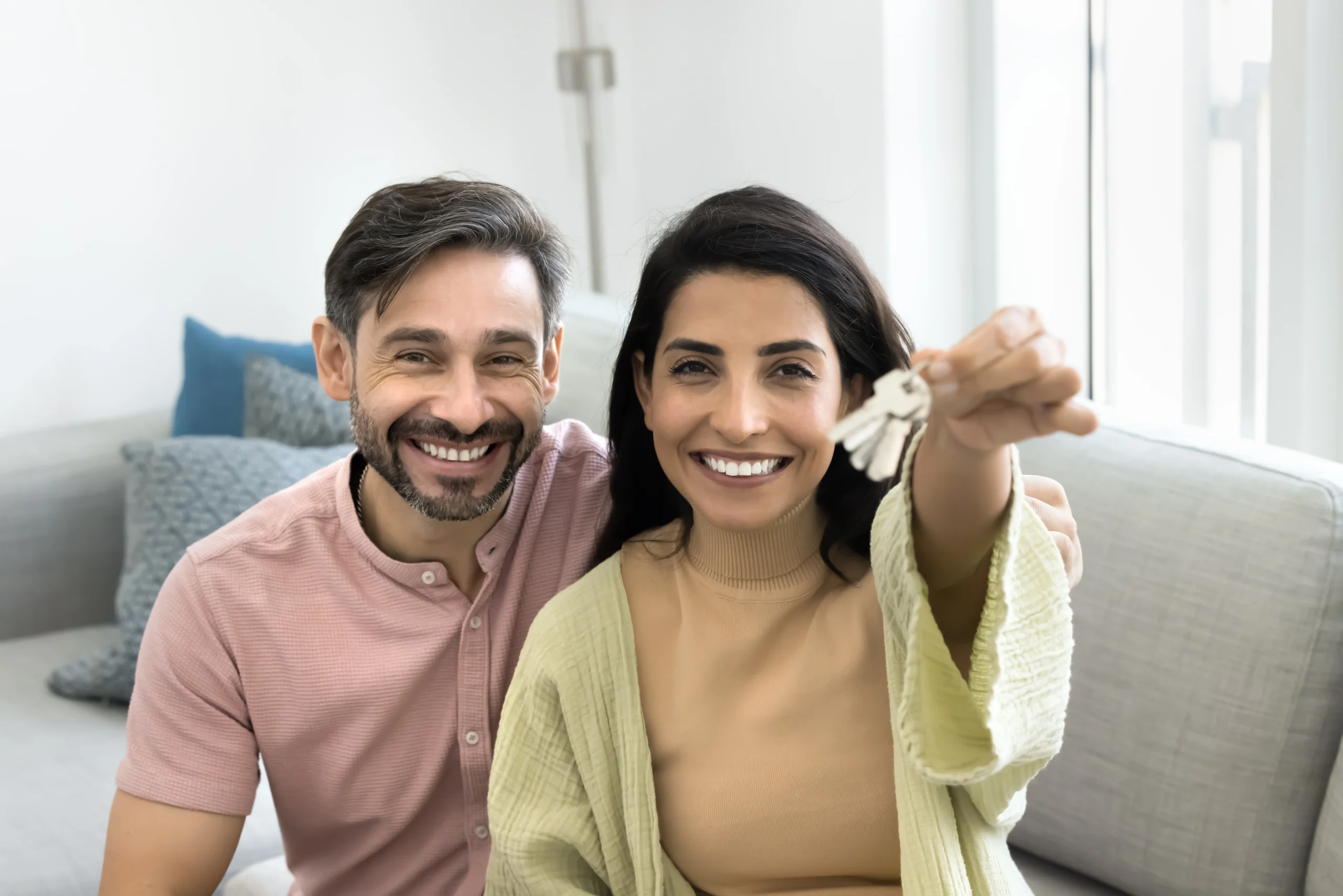A happy couple smile on the couch holding up the keys to the new home they've bought