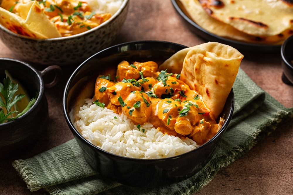 A black bowl of creamy coconut curry sits on one side, paired with fluffy white rice on the other. A piece of naan is tucked in, garnished with fresh cilantro. The scene is warm and inviting, with a green napkin underneath.