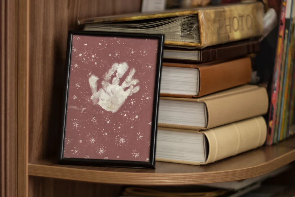 a toddler-size hand print in white paint on dark red paper surrounded by painted white stars in a black frame on a bookshelf