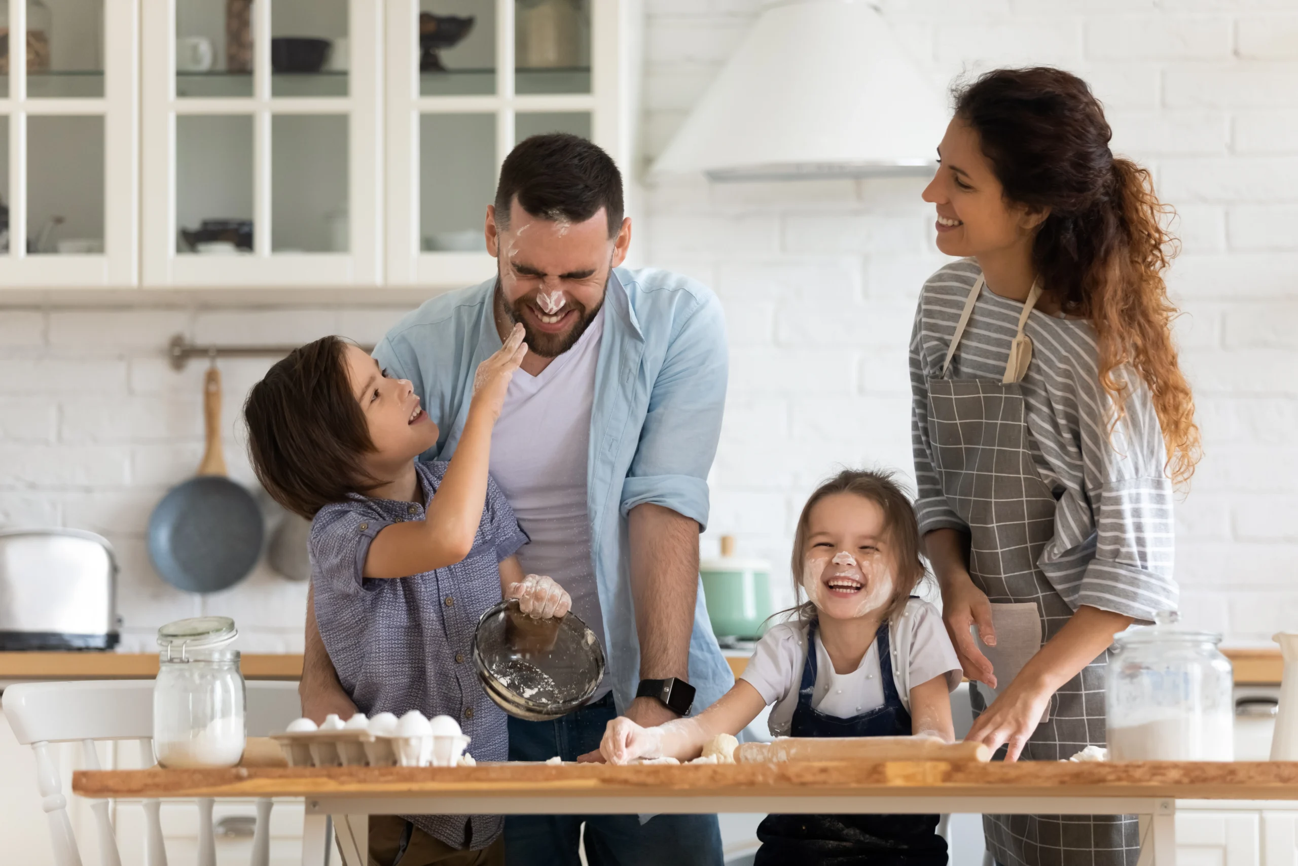 A mum, dad, and two young kids bake together at the kitchen island, laughing as one of the kids has smeared flower on his dad