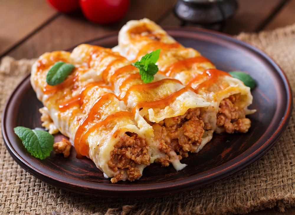 Close-up of baked enchiladas on a dark plate, with browned cheese and tomato sauce, garnished with fresh mint leaves. Cozy and appetizing feel.