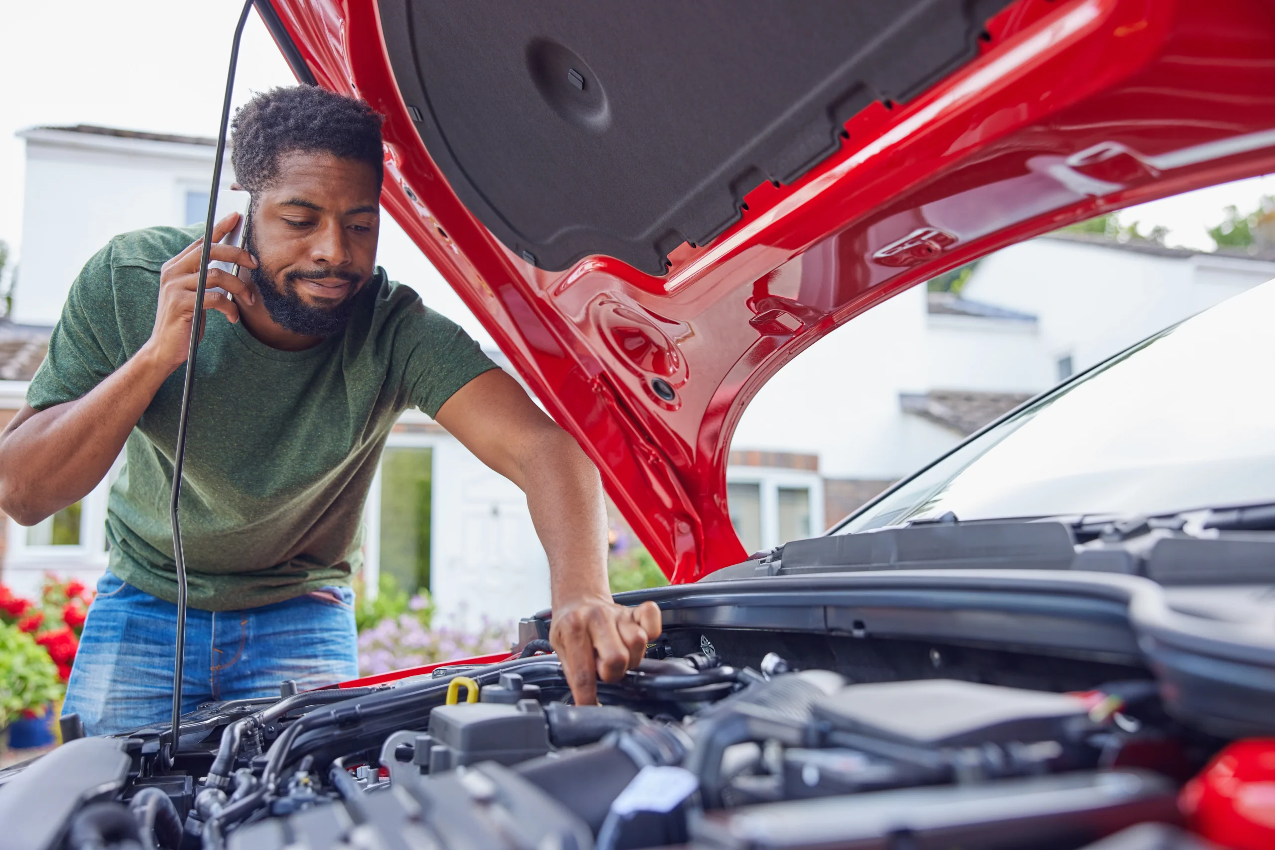 A man leans into the open hood of his car while on the phone to a mechanic, looking frustrated
