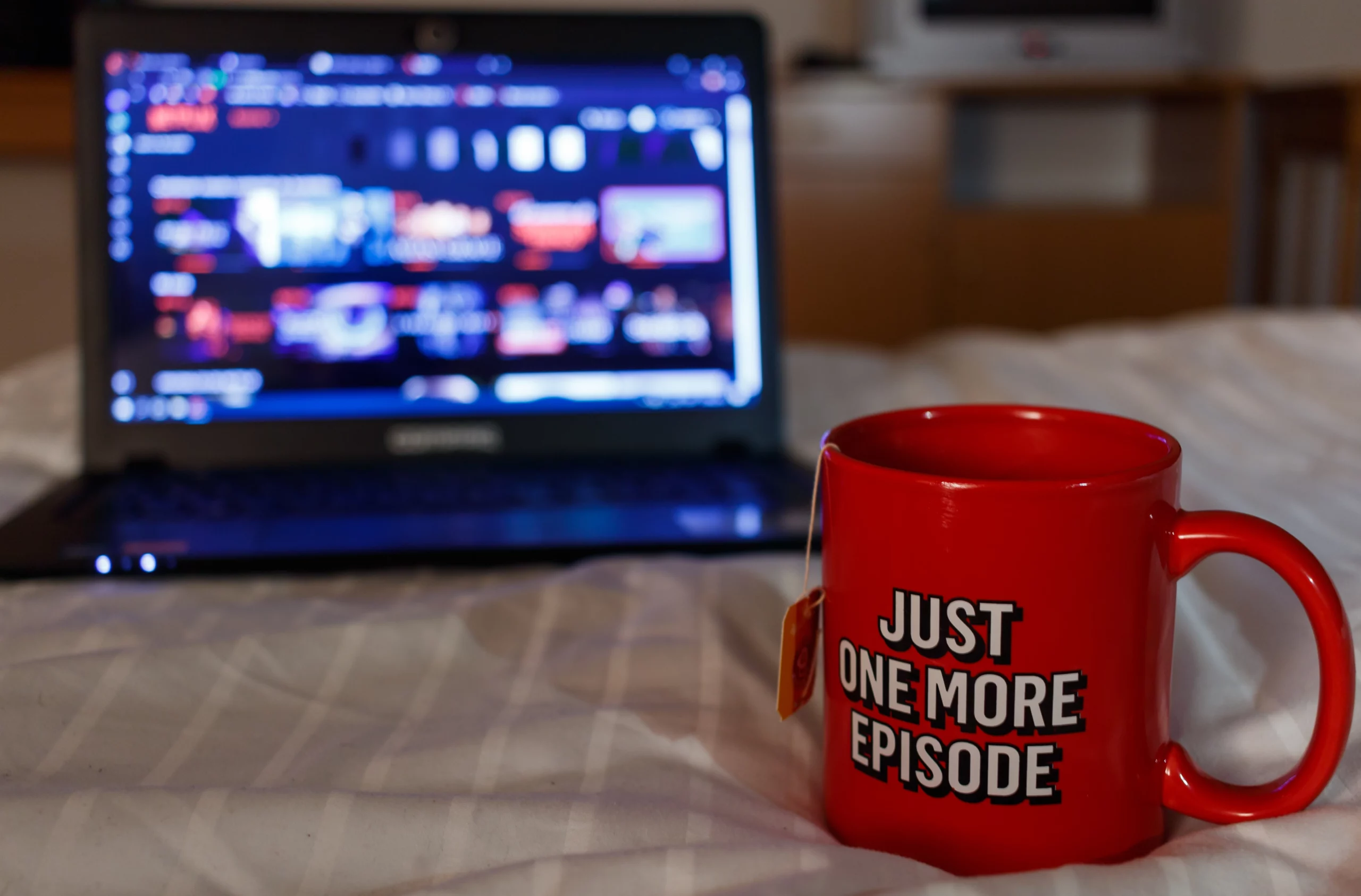 Laptop showing blurred free streaming service on a bed with mug with "Just one more episode" lettering resting in front with tea bag