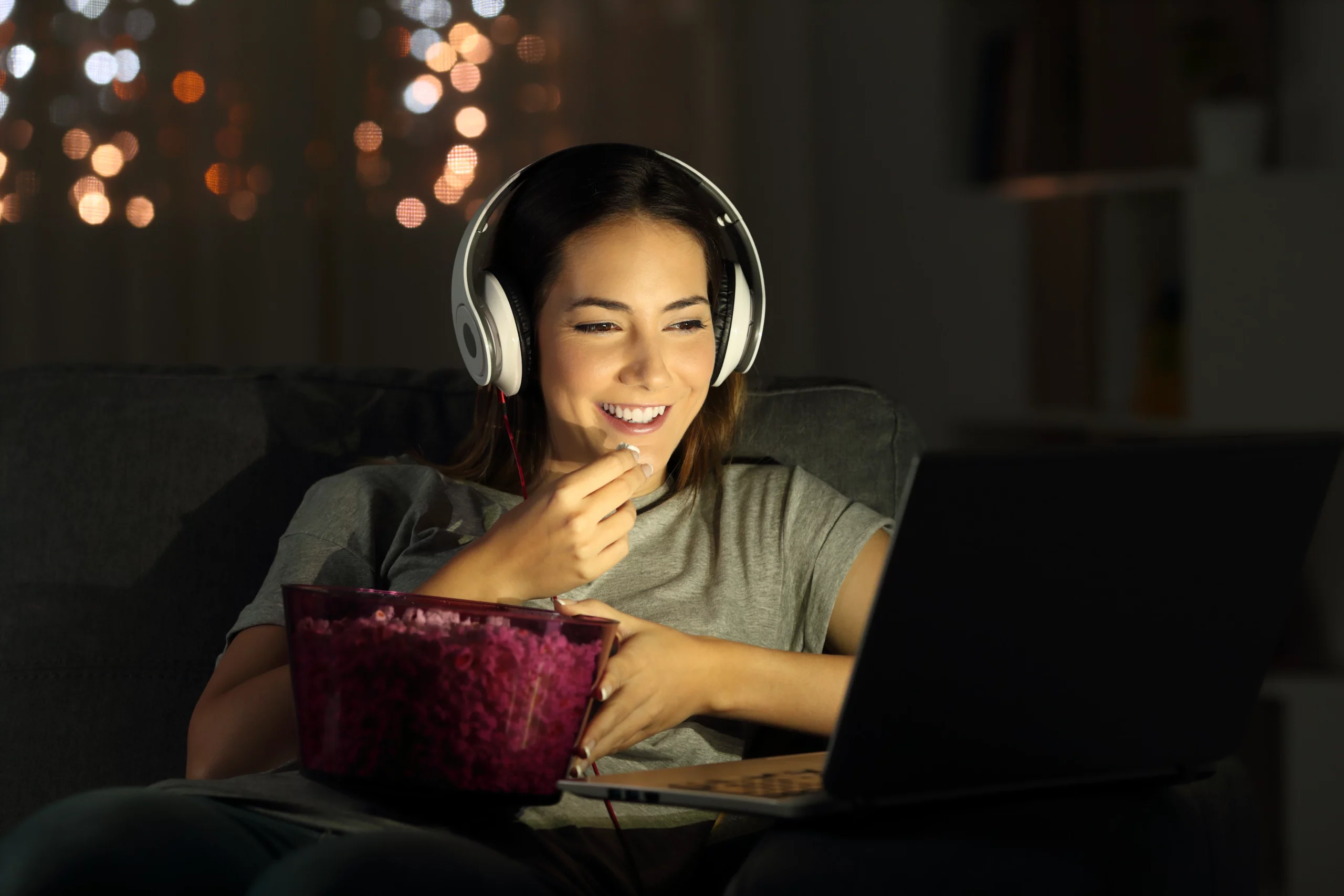 woman watching free-to-air TV on her laptop on a couch with headphones while eating from a pink bowl of popcorn