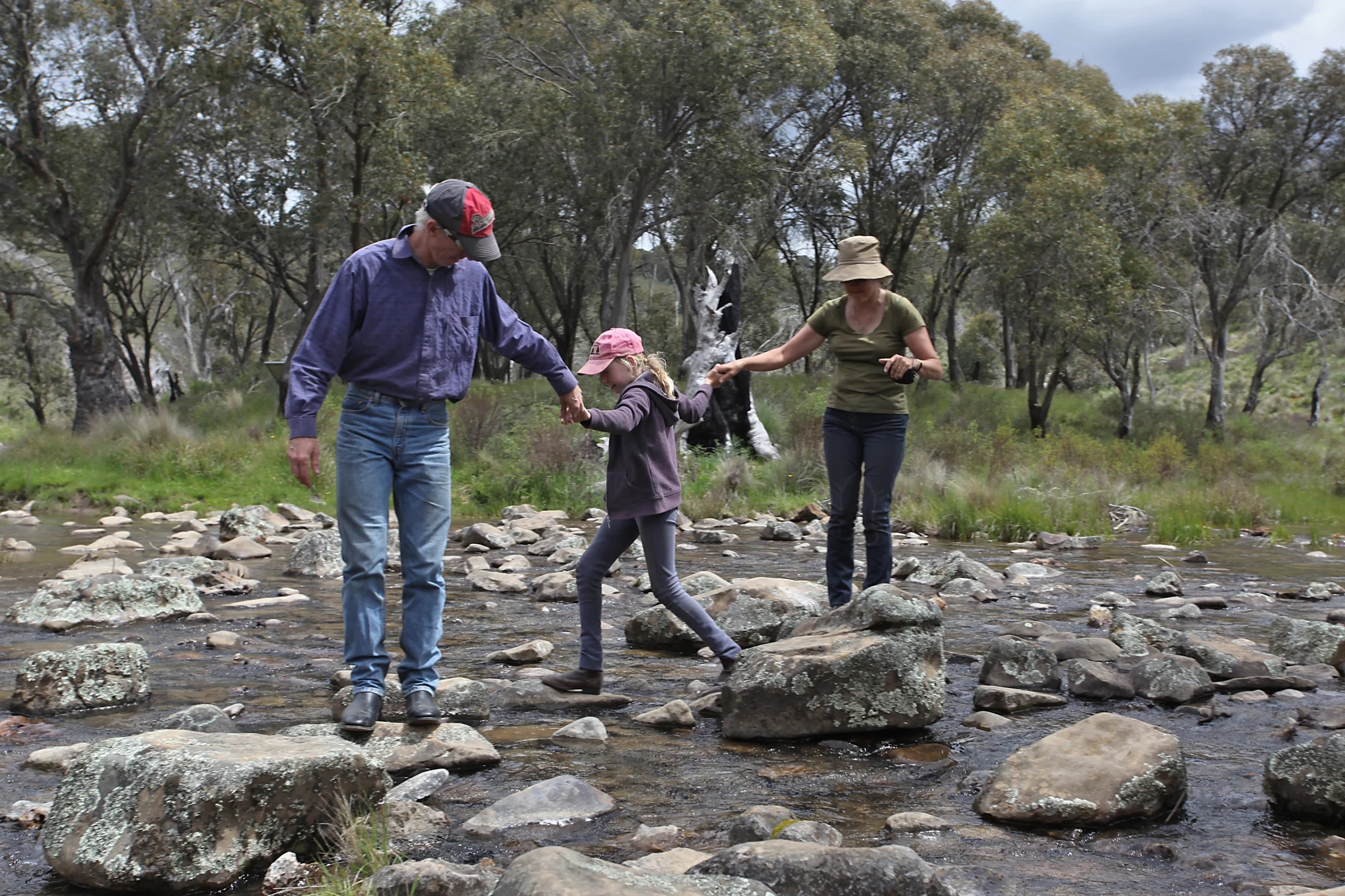 Grandma and grandpa on a hike, hopping rocks over a creek with their granddaughter