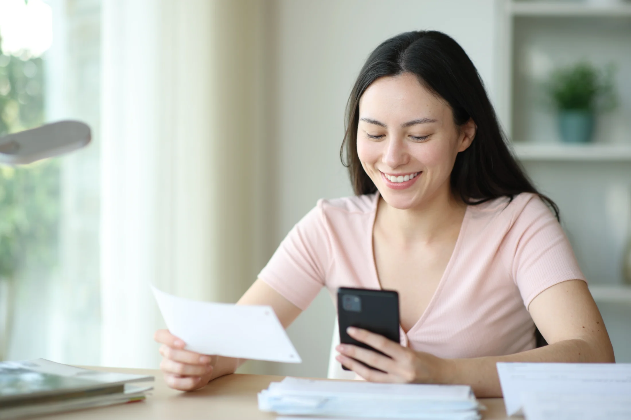 Smiling woman sits at a table holding her phone and a slip of paper as she works out how to fix her credit rating