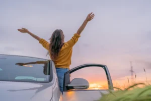 A woman leans out the drivers side of her car facing the sunset with her hands in the air