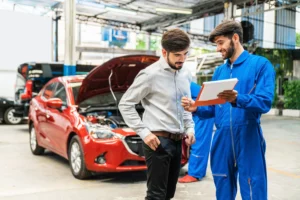 A mechanic standing in front of a red car with the hood open shows a man a checklist of repairs