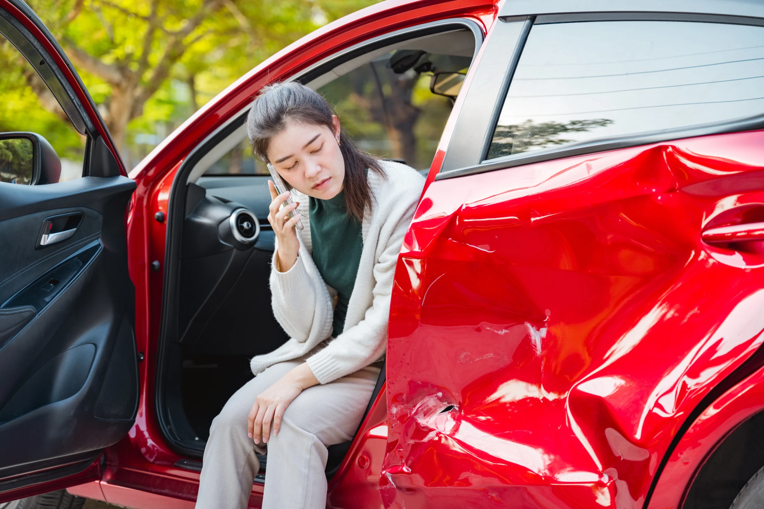 A woman sits in a red car with crash damage calling her insurance