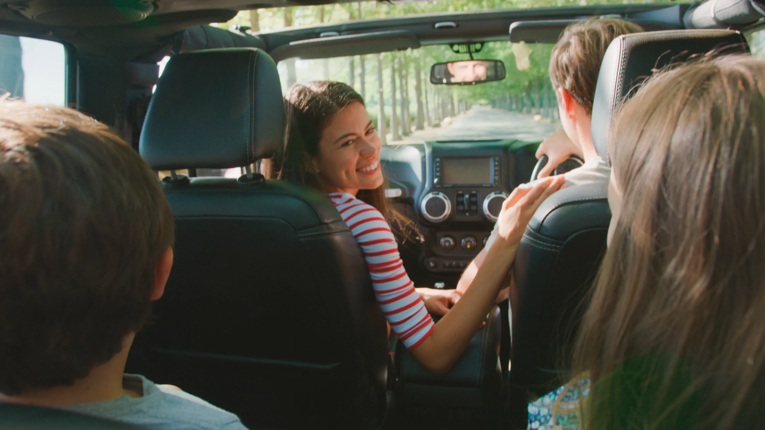 A mother turns and looks at her kids in the backseat of a car driving down an tree-lined road