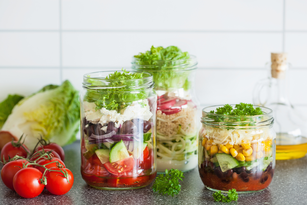 Three glass jars filled with Lentil salad, layered with vegetables, cheese, and greens, are displayed on a countertop—perfect vegetarian lunch ideas to take to work—with tomatoes, lettuce, and an oil bottle in the background.