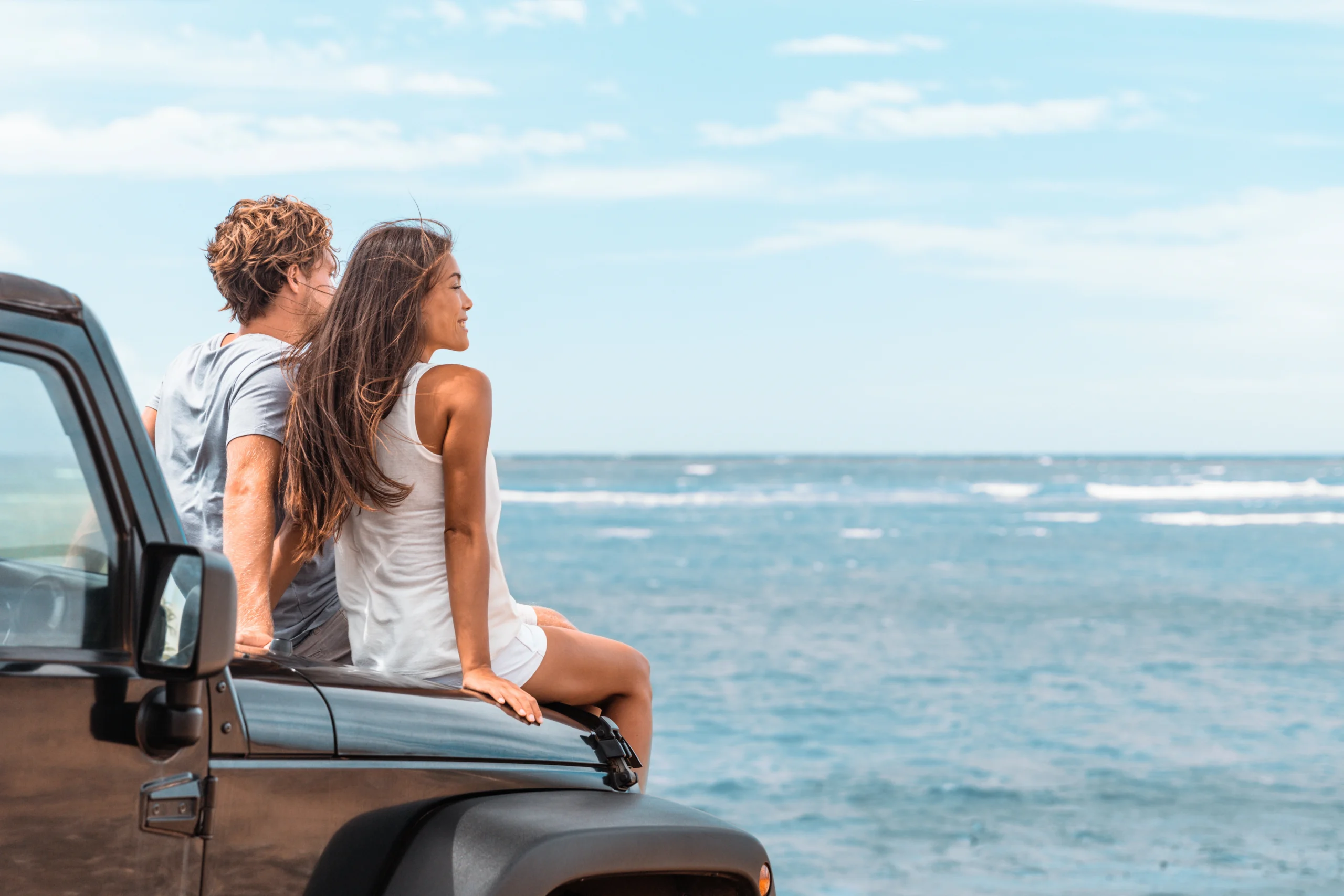Smiling couple sits on the bonnet of their car looking out at the ocean