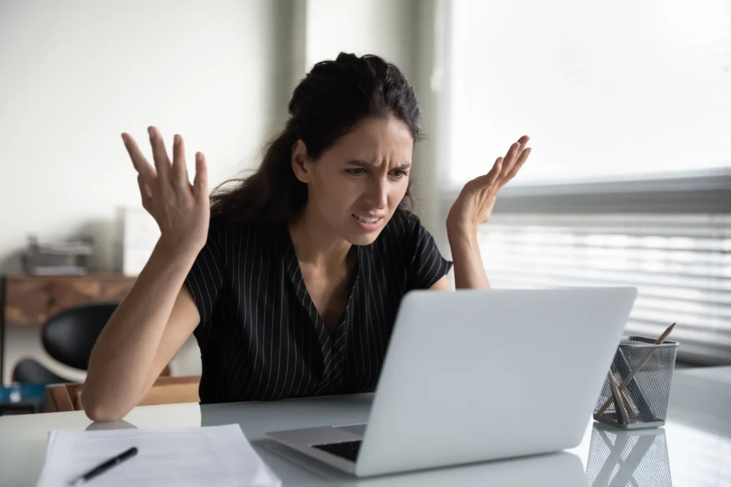 Woman at a desk looks frustrated, raising her hands while staring at a laptop. Papers and a pencil holder are nearby, conveying frustration at work.
