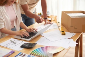 A couple at a desk full of colour swatches, building plans, and budgets looking for unexpected renovation costs