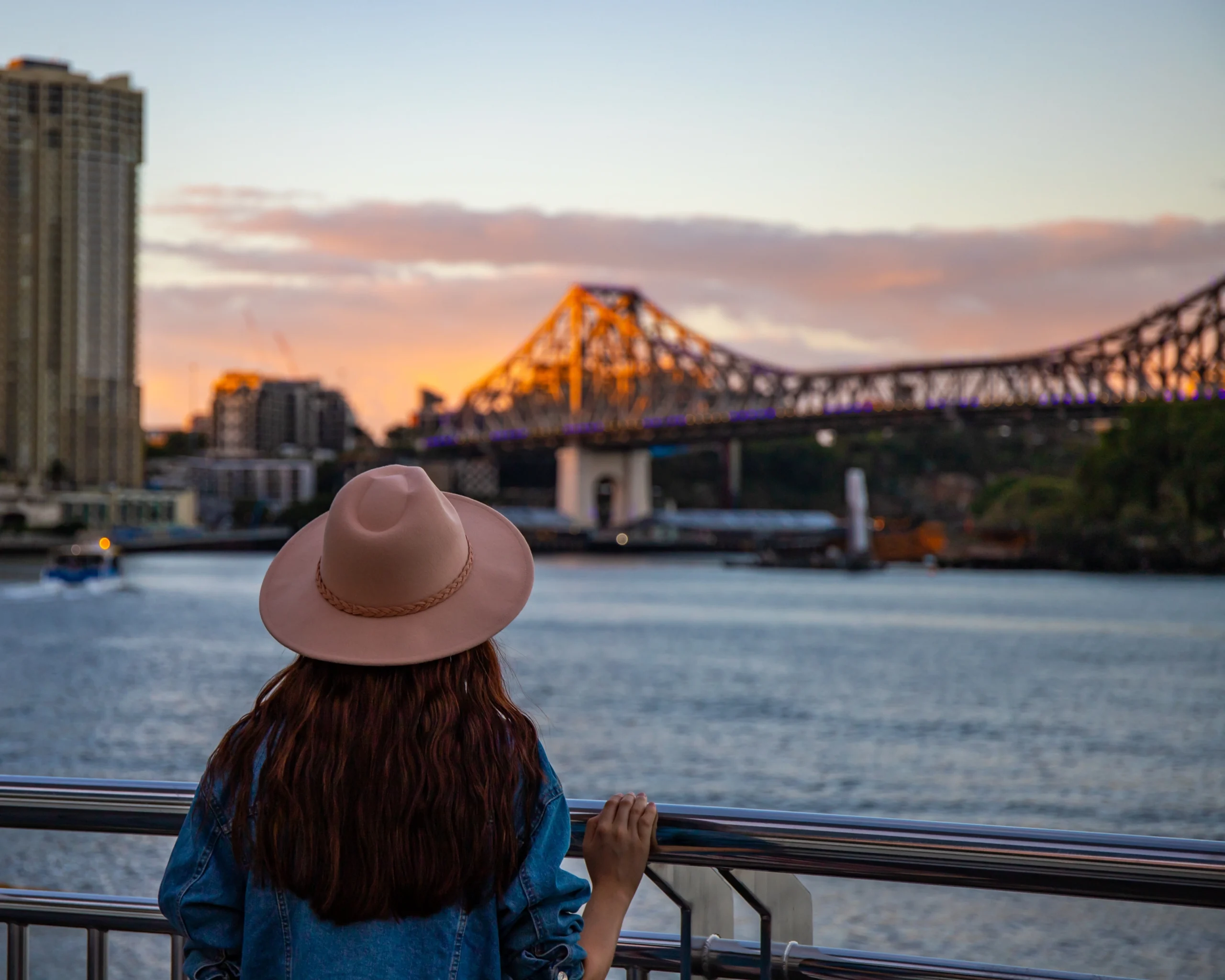 A woman wearing a broad rim hat stares out at the Brisbane River with the Storey Bridge in the background