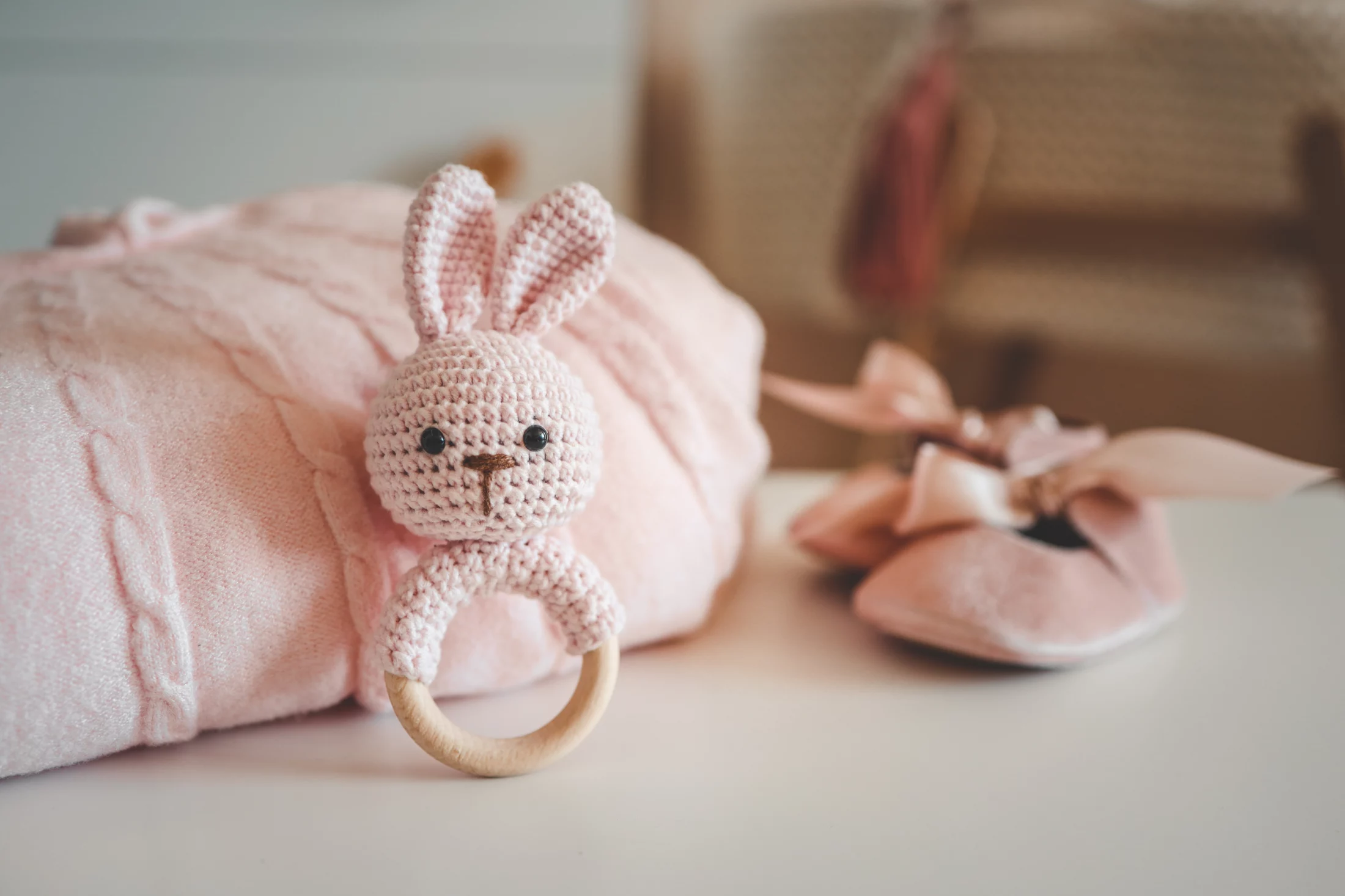 Soft pink baby blanket with a crocheted bunny teether and matching bow shoes arranged on a nursery shelf.