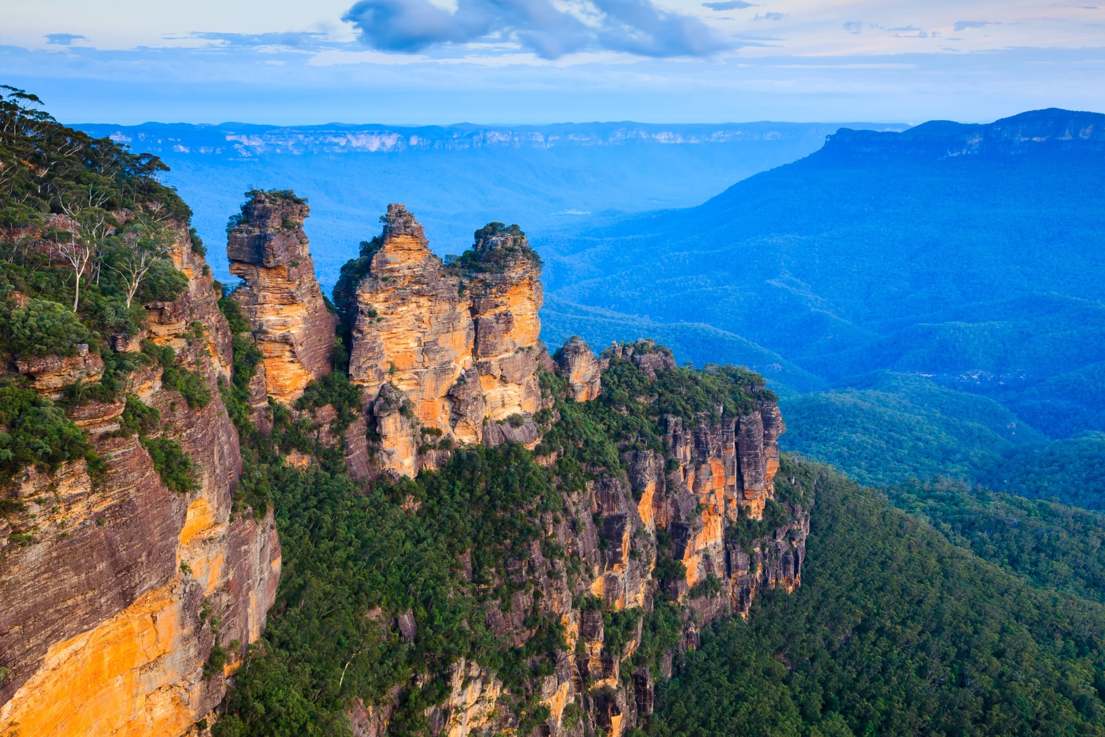 The Three Sisters rock formation in the Blue Mountains, Australia, with expansive mountain views and a partly cloudy sky.