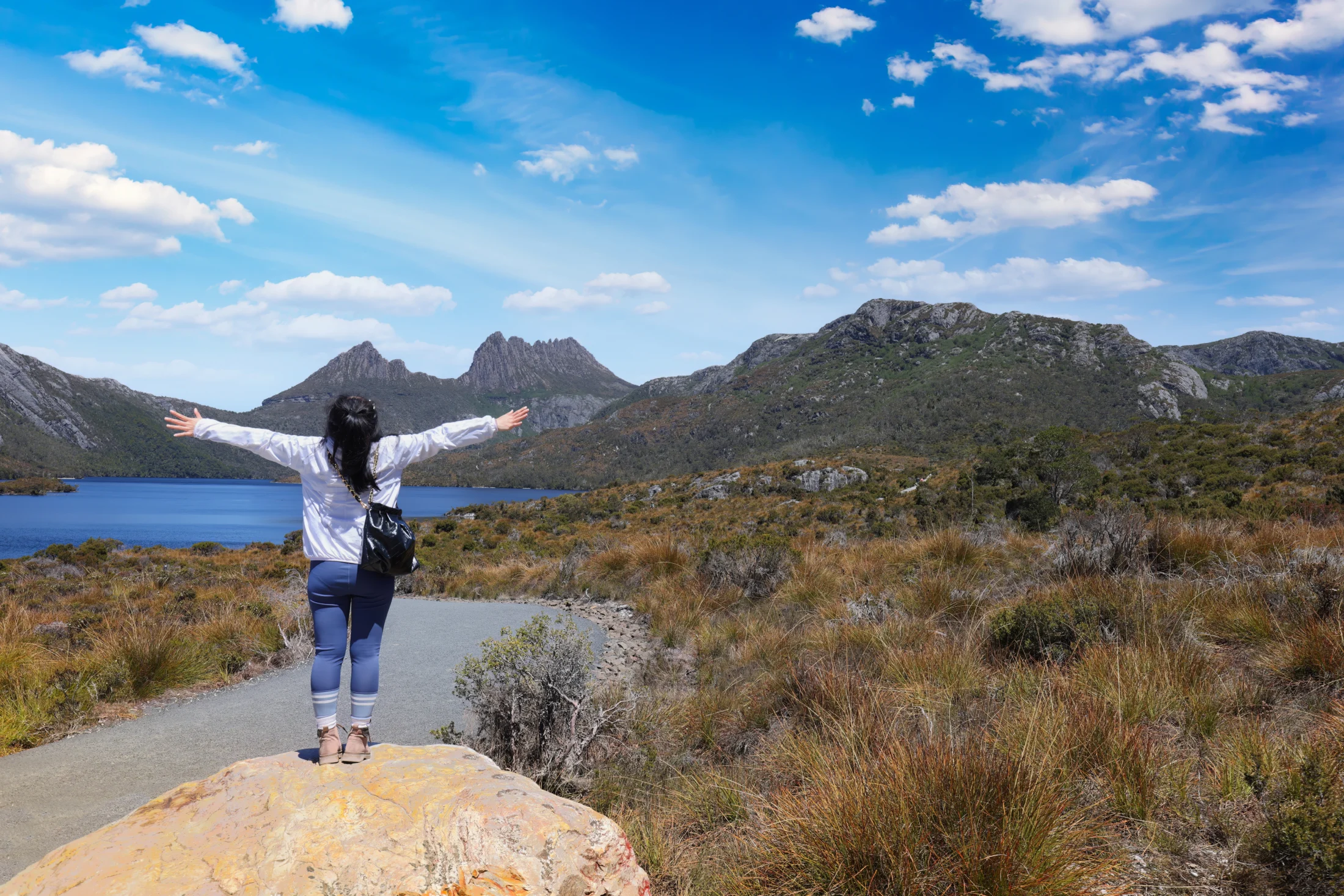 The image depicts a person standing on a large rock, arms outstretched, facing the expansive natural landscape at Cradle Mountain, Tasmania.