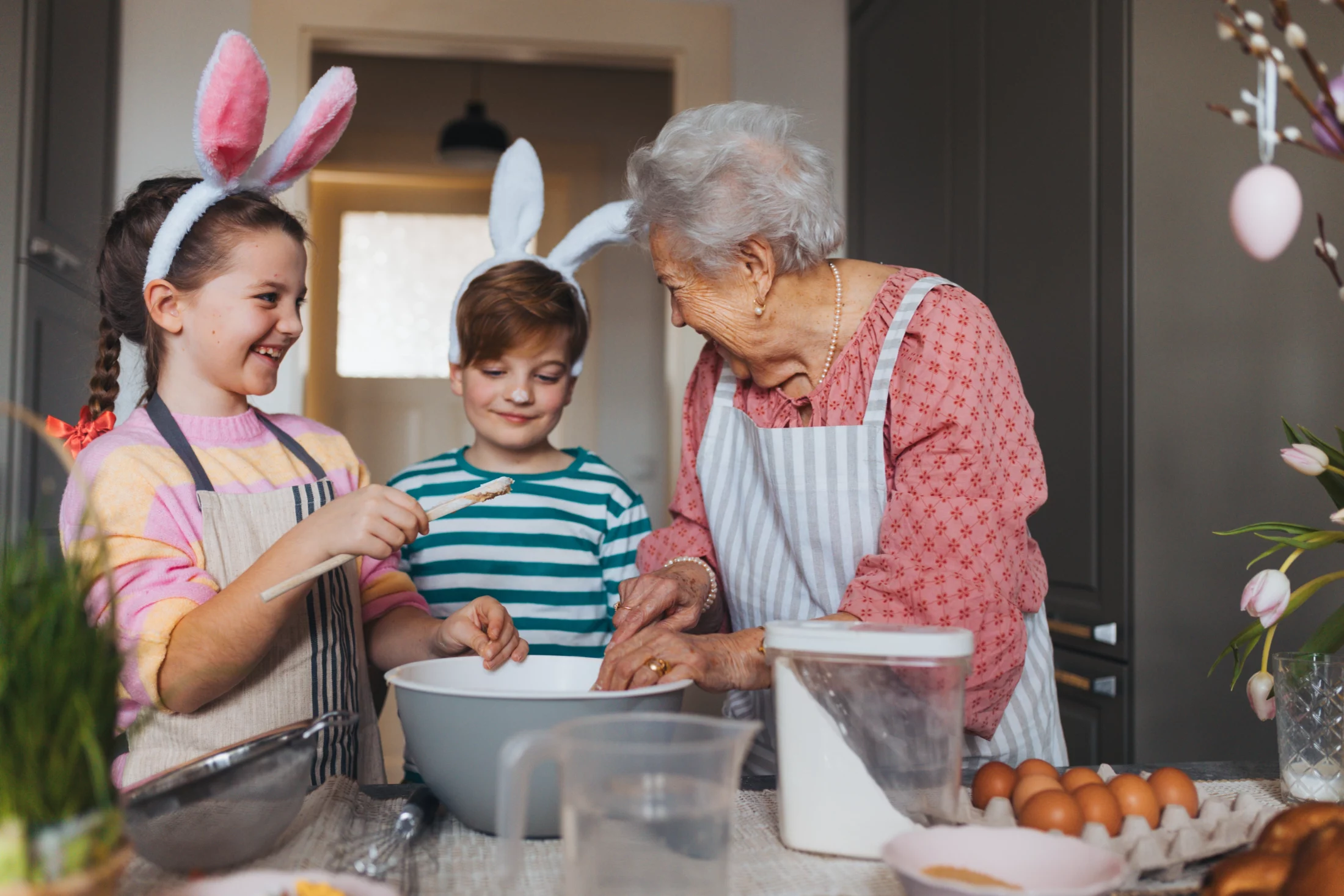 Grandmother and two children wearing bunny ears and aprons baking together for Easter in a bright kitchen with eggs and mixing bowls on the counter.