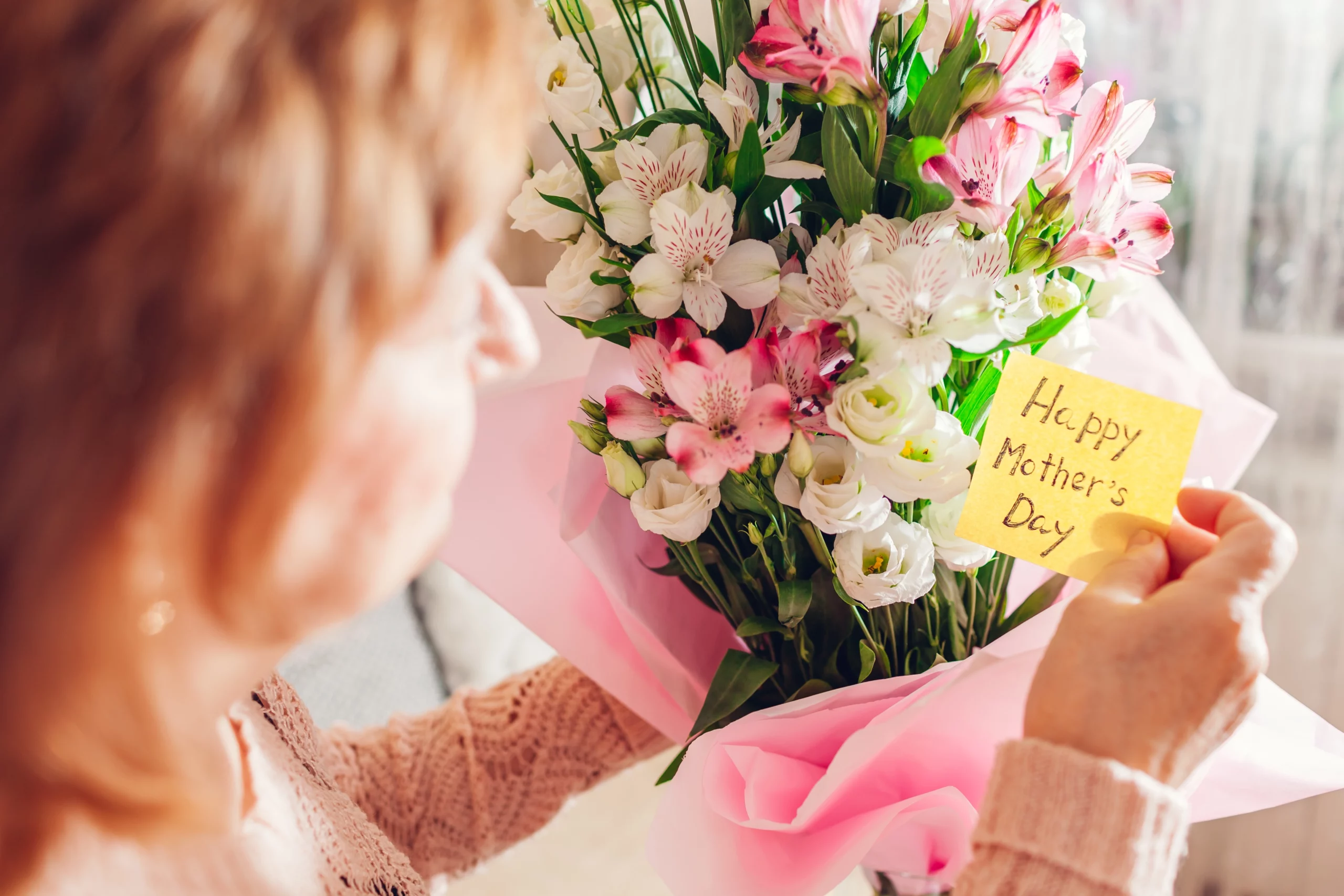 Mother holding a bouquet of pink and white flowers in light pink wrapping and a note that says "Happy Mother's Day"