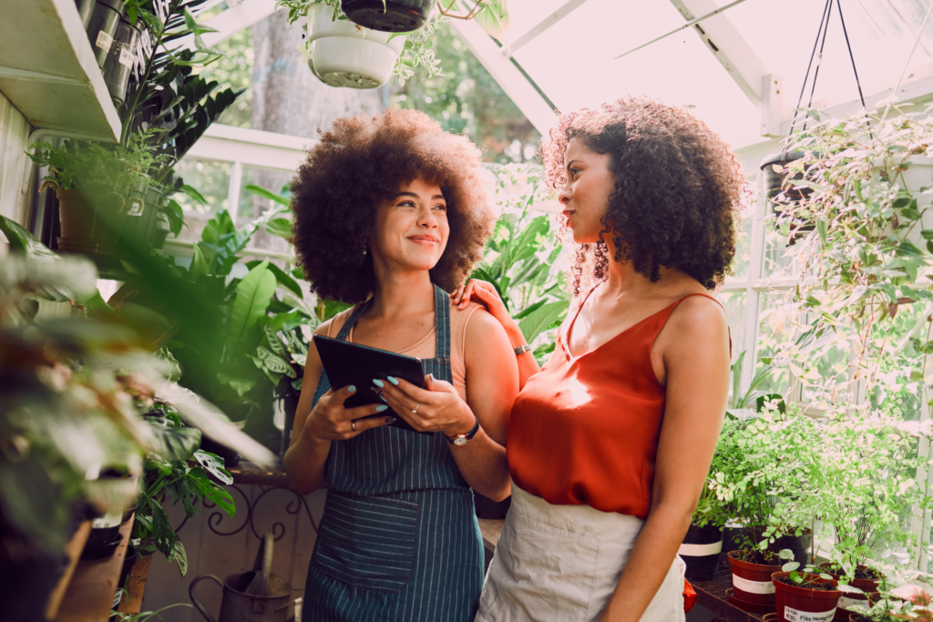 Two women look at a tablet together while standing inside a greenhouse