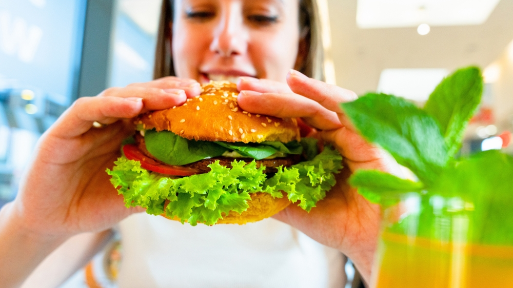 A woman smiles while holding a large salmon burger with lettuce, tomato, spinach, and a sesame seed bun. A mint garnish is next to a glass with orange liquid.