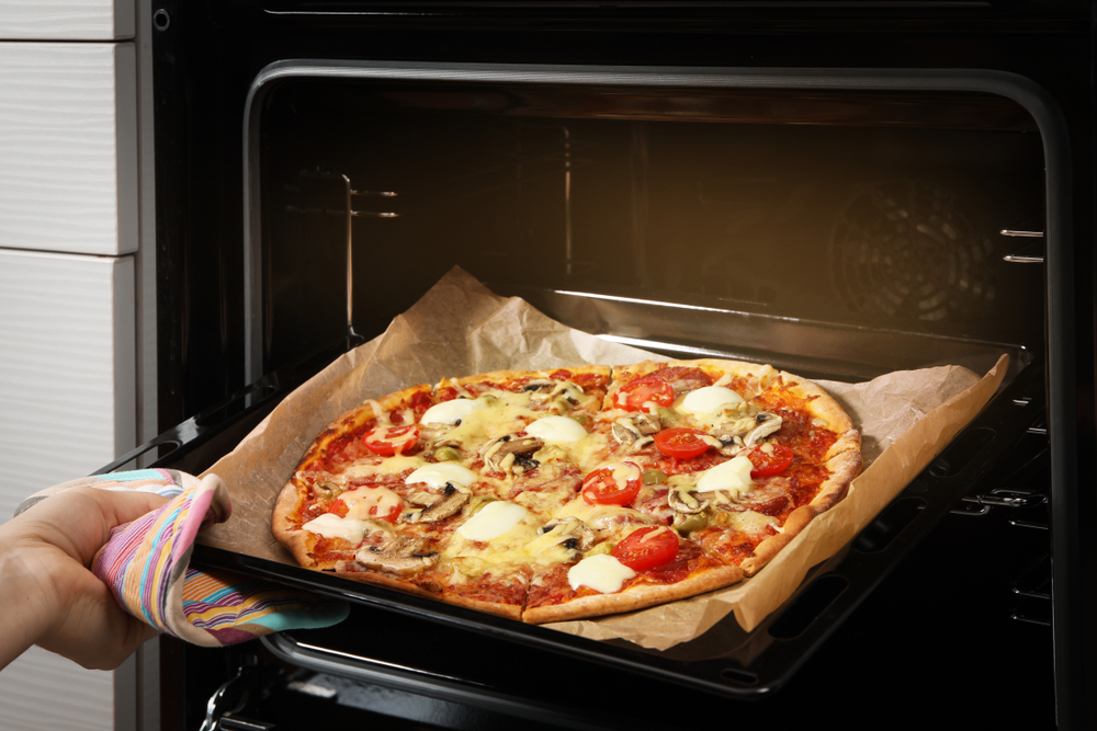 A freshly baked pizza with tomatoes and cheese is being taken out of an oven on a baking tray lined with parchment paper. A hand with an oven mitt holds the tray.