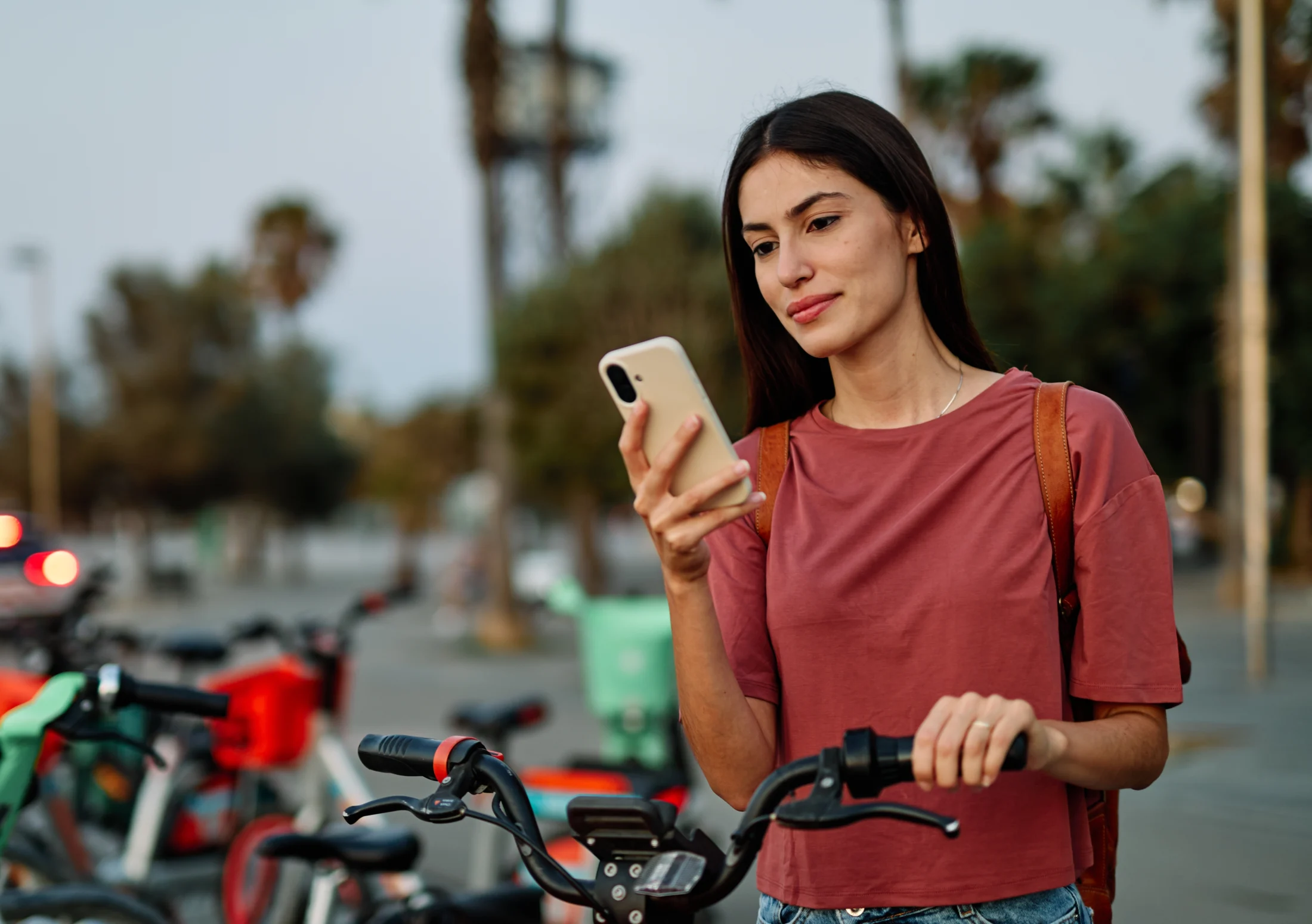 Woman standing outdoors holding a bicycle and checking her phone for emergency loan options, with rental bikes and palm trees in the background.
