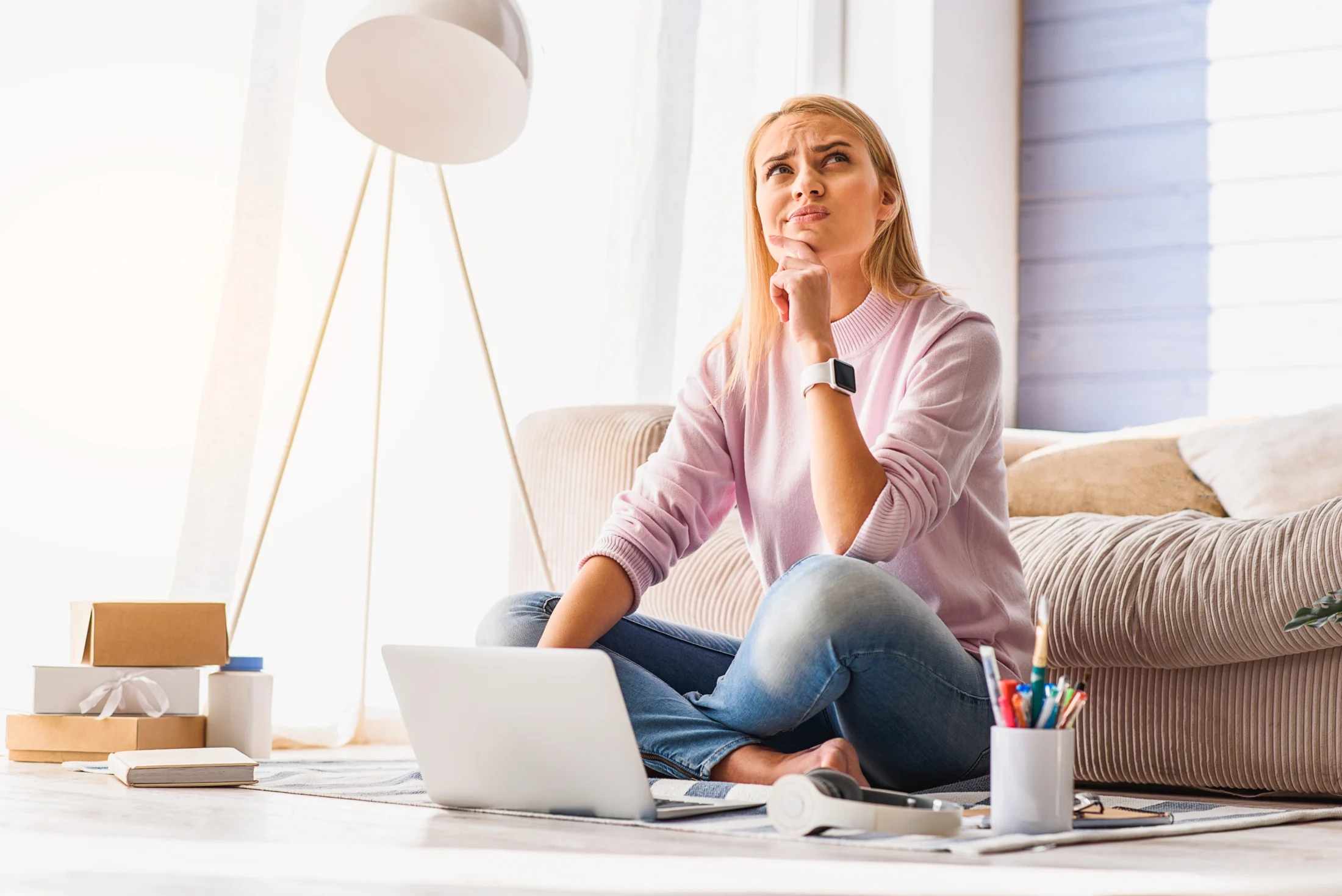 A thoughtful woman in a pink sweater sits cross-legged in a cozy room, using a laptop. She gazes upward, surrounded by books and a cup of pens.