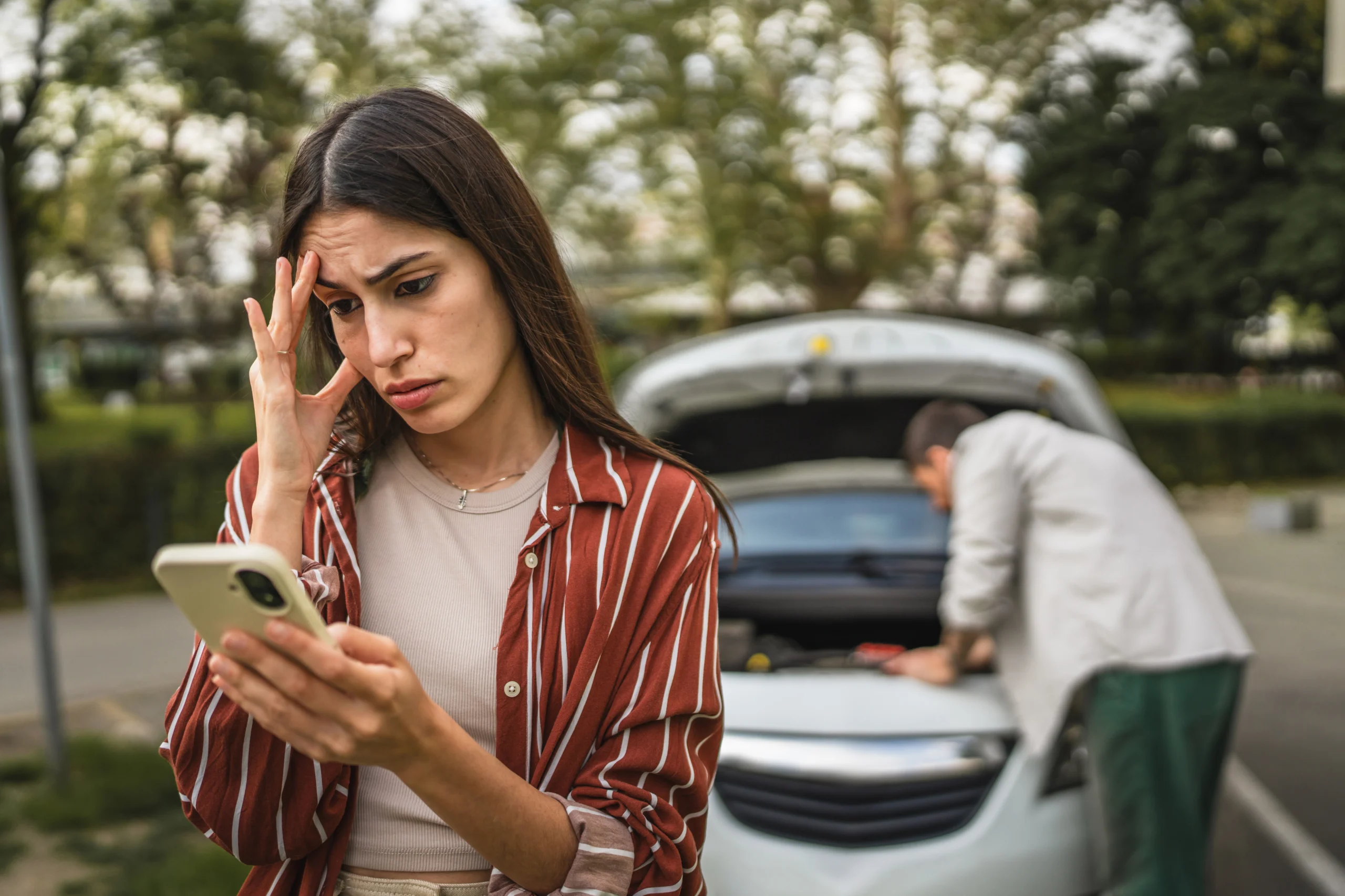 A worried woman looks at her phone, standing beside a car with an open hood. In the background, a person inspects the engine. The setting is outdoors.